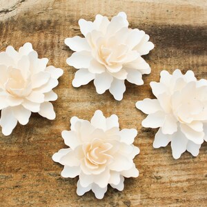 May include: Four white paper flowers with a layered, ruffled design. The flowers are arranged on a rustic wooden background.