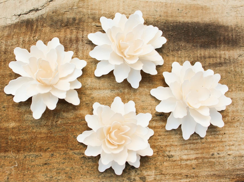 May include: Four white paper flowers with a layered, ruffled design. The flowers are arranged on a rustic wooden background.
