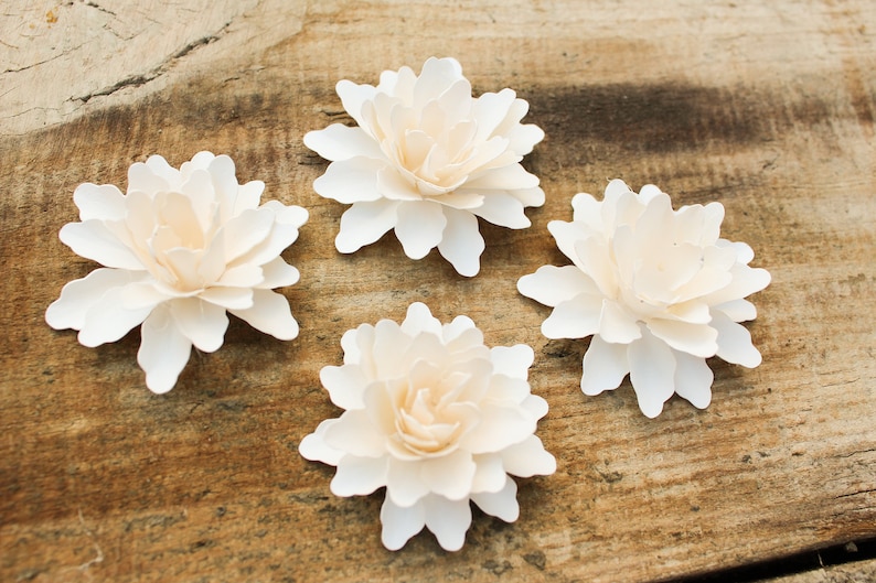 May include: Four white paper flowers with a layered, ruffled design. The flowers are arranged on a wooden surface.