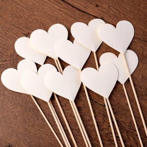 May include: A group of white heart-shaped paper decorations on wooden sticks. The hearts are fanned out against a brown wooden background. Suitable for crafting or party decorations.