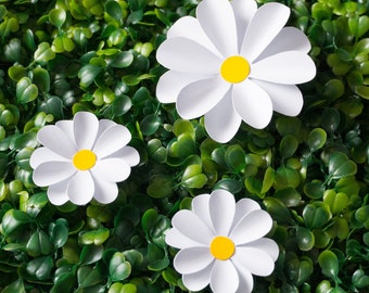 May include: Three white paper daisies with yellow centers are arranged on a green artificial grass background.
