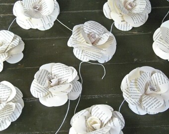 Peut inclure: Une guirlande de fleurs en papier blanc faites à partir de pages de livres. Les fleurs sont enfilées sur un fil blanc et sont suspendues à une surface en bois vert foncé.