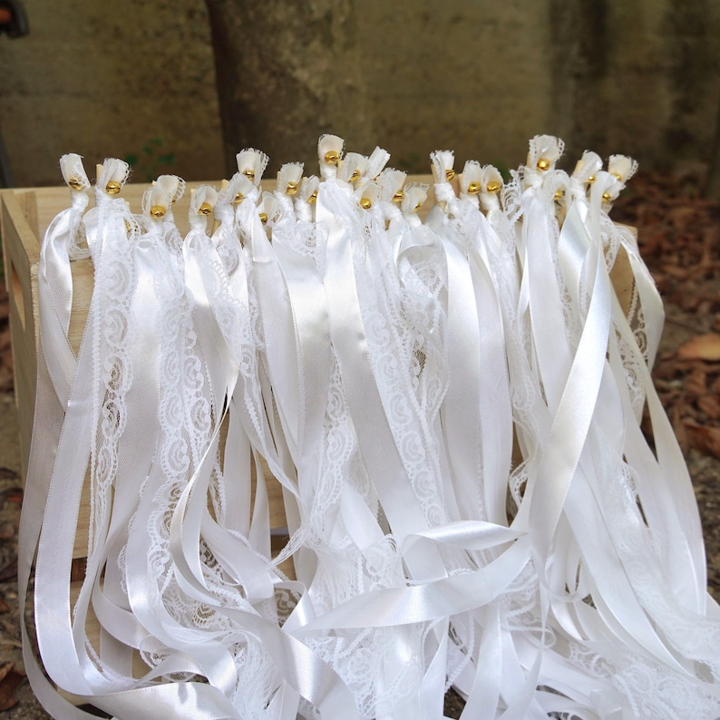 May include: A collection of white ribbons, some with delicate lace trim, hang from a wooden structure. Each ribbon is topped with a small gold bead. The ribbons are of varying widths and textures, creating a layered effect.