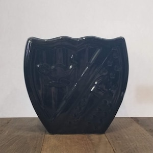 May include: A dark blue ceramic vase shaped like a shield with a raised design of two crossed swords and a bird.