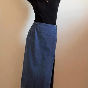 May include: A long denim skirt with a side slit. The skirt is a dark blue color and has a straight fit.