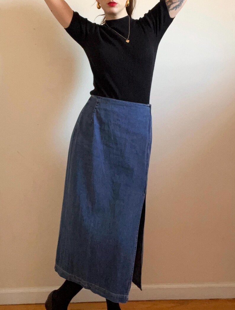 May include: A long denim skirt with a side slit. The skirt is a dark blue color and has a straight fit.