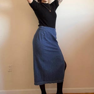 May include: A long denim skirt with a slit on the side. The skirt is a dark blue color and is worn with a black mock neck shirt.