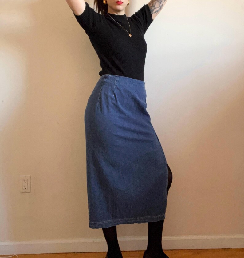 May include: A long denim skirt with a slit on the side. The skirt is a dark blue color and is worn with a black mock neck shirt.