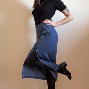 May include: A woman wearing a black mock neck shirt and a blue denim skirt with a slit up the side. She is wearing black tights and black heeled boots.