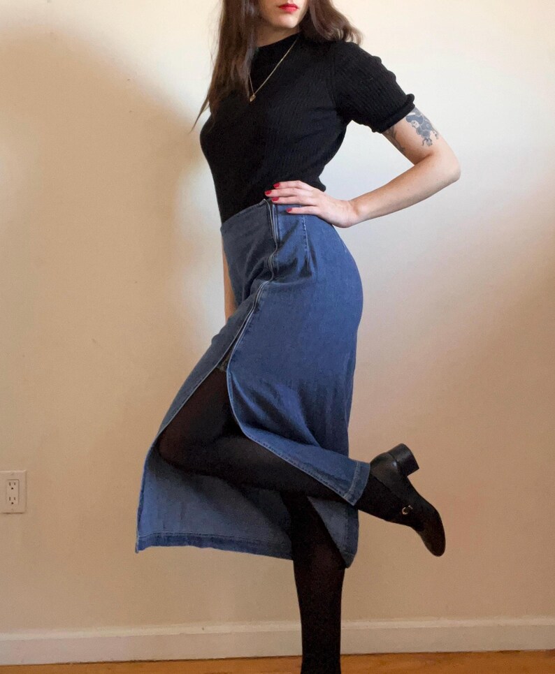 May include: A woman wearing a black mock neck shirt and a blue denim skirt with a slit up the side. She is wearing black tights and black heeled boots.
