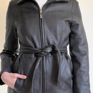 May include: A black leather jacket featuring a full-length front zipper, a pointed collar, and long sleeves. The jacket includes a matching black leather tie belt at the waist and two side pockets. The material has a smooth, matte finish.