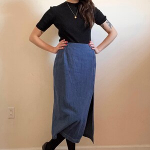 May include: A woman wearing a black mock neck shirt and a long blue denim skirt with a side slit.