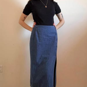 May include: A long denim skirt with a side slit. The skirt is a dark blue color and has a straight fit. The skirt is worn with a black mock neck shirt.