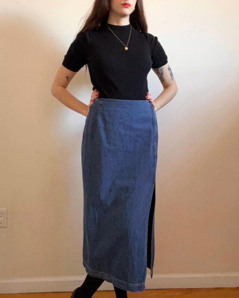 May include: A long denim skirt with a side slit. The skirt is a dark blue color and has a straight fit. The skirt is worn with a black mock neck shirt.