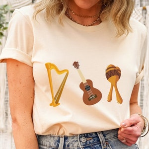 May include: A beige t-shirt with a graphic of a harp, ukulele, and maracas. The instruments are brown, yellow, and orange.