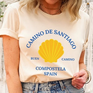 May include: A cream colored t-shirt with a blue and yellow graphic design. The graphic features a scallop shell with the text "Camino de Santiago", "Buen Camino", "Compostela", and "Spain".