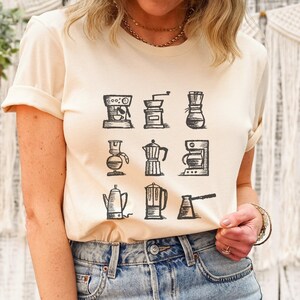 May include: A beige t-shirt with a graphic of nine different coffee makers in black and white. The coffee makers include a French press, a percolator, a drip coffee maker, and a pour-over coffee maker.
