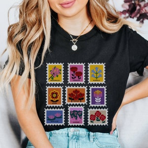 May include: Black t-shirt featuring a graphic of nine colourful flower stamps. Each stamp depicts a different flower with its name printed below: Bougainvillea, Carnation, Bluebells, Gardenia, Poppies, Lantana, Water Lilies, Geraniums, and Iceland Poppy.