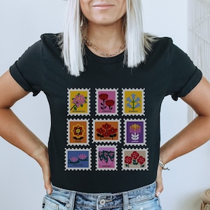 May include: Black t-shirt featuring a graphic of nine colourful flower illustrations in the shape of postage stamps. Each stamp depicts a different flower with its name printed below: Bougainvillea, Carnation, Bluebells, Gazania, Poppies, Lantana, Water Lilies, Geranium, and Dianthus.