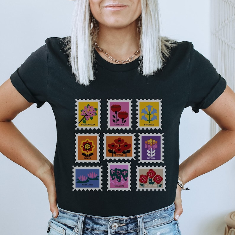 May include: Black t-shirt featuring a graphic of nine colourful flower illustrations in the shape of postage stamps. Each stamp depicts a different flower with its name printed below: Bougainvillea, Carnation, Bluebells, Gazania, Poppies, Lantana, Water Lilies, Geranium, and Dianthus.