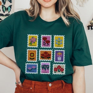 May include: A dark green t-shirt featuring a vintage-style floral print. The print includes nine different flowers, each in a separate square frame with a white border. The flowers are labelled with their names, including Bougainvillea, Carnation, Bluebells, Gazania, Poppies, Lantana, Water Lilies,  Exacum, and Valerianca.