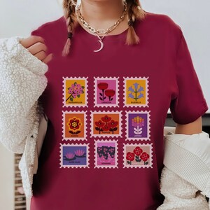 May include: A maroon t-shirt featuring a graphic of nine colourful flower stamps. Each stamp depicts a different flower with its name printed below: Bougainvillea, Carnation, Bluebells, Gazania, Poppies, Lantana, Water Lilies, Geraniums, and Valerian Root.