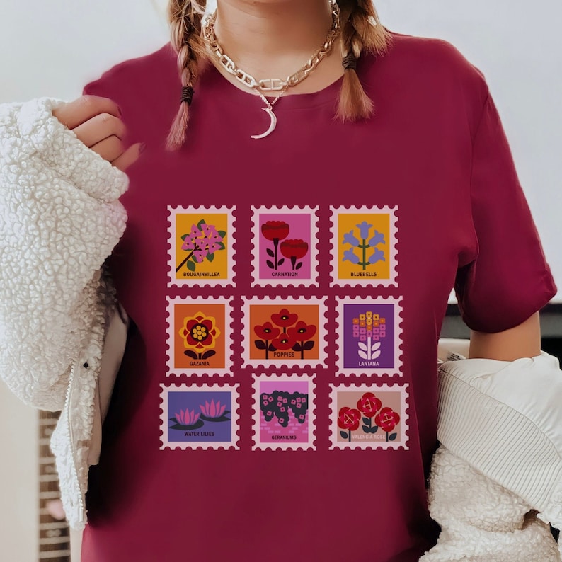 May include: A maroon t-shirt featuring a graphic of nine colourful flower stamps. Each stamp depicts a different flower with its name printed below: Bougainvillea, Carnation, Bluebells, Gazania, Poppies, Lantana, Water Lilies, Geraniums, and Valerian Root.
