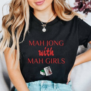 May include: Black t-shirt with the text "Mahjong with Mah Girls" in red lettering. A green and white mahjong tile is pictured below the text.