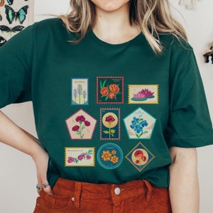 May include: A green t-shirt with a vintage-style design featuring a collection of colorful flower illustrations on postage stamps. The stamps feature various flowers, including bluebells, peonies, water lilies, carnations, roses, forget-me-nots, wildflowers, and daisies.