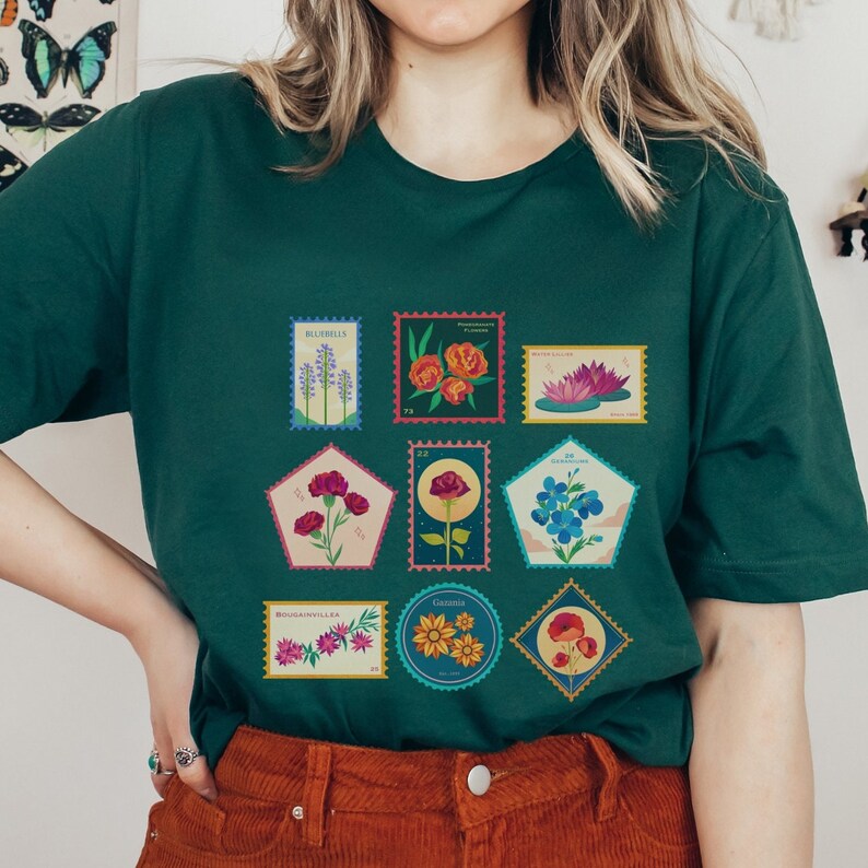 May include: A green t-shirt with a vintage-style design featuring a collection of colorful flower illustrations on postage stamps. The stamps feature various flowers, including bluebells, peonies, water lilies, carnations, roses, forget-me-nots, wildflowers, and daisies.