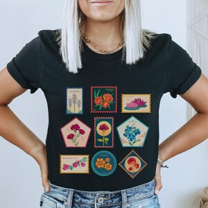 May include: Black t-shirt with a graphic of nine colorful flower stamps. Each stamp features a different flower and the name of the flower. The stamps are arranged in a grid pattern.