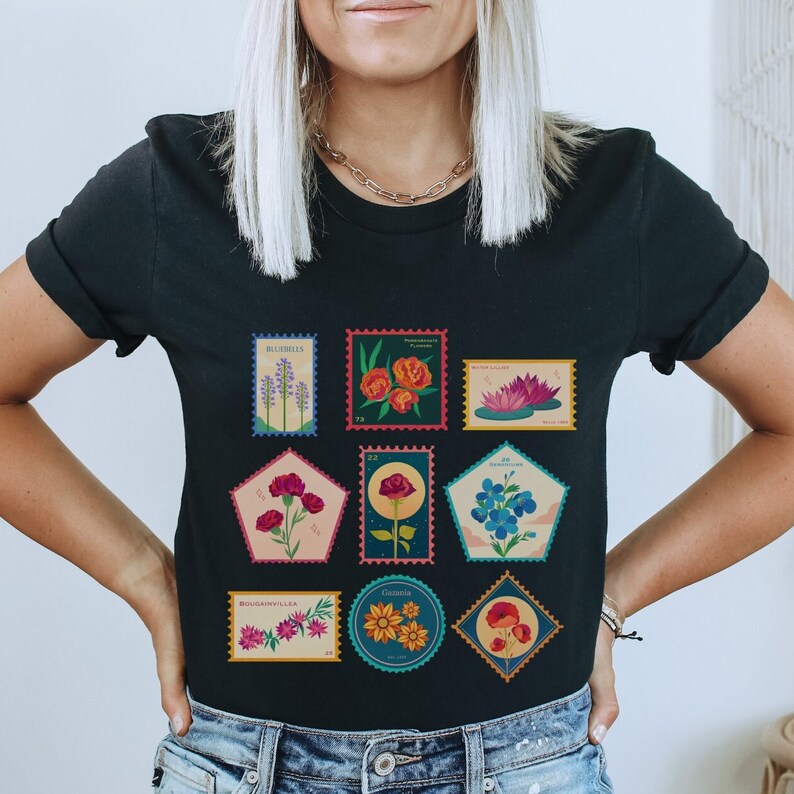 May include: Black t-shirt with a graphic of nine colorful flower stamps. Each stamp features a different flower and the name of the flower. The stamps are arranged in a grid pattern.