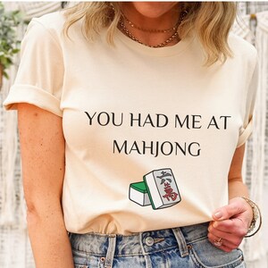 May include: A cream colored t-shirt with the text "You had me at Mahjong" and a graphic of two mahjong tiles.