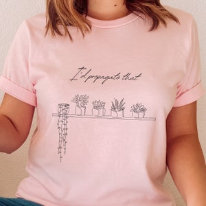 May include: A pink t-shirt with the text "I'd propagate that" and a line drawing of potted plants on a shelf.