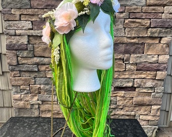 May include: A green and yellow yarn wig with a floral crown on a white mannequin head. The crown has pink and white flowers and green leaves.