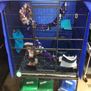 May include: A black metal cage with a blue fabric cover. The cage has multiple levels and is filled with colorful toys and bedding. The cage is on a stand with two shelves, one with green and one with blue fabric.