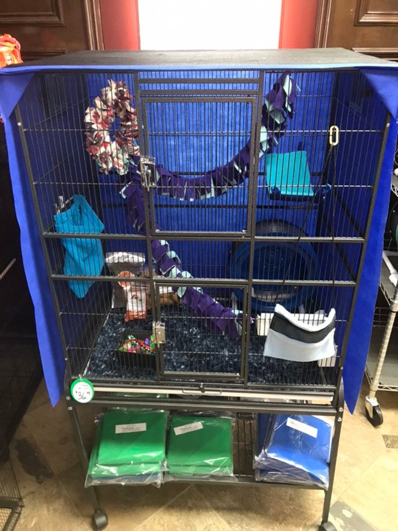 May include: A black metal cage with a blue fabric cover. The cage has multiple levels and is filled with colorful toys and bedding. The cage is on a stand with two shelves, one with green and one with blue fabric.