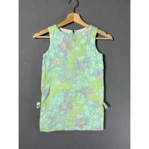 May include: A sleeveless dress with a floral pattern in shades of green, purple, and yellow. The dress is hanging on a wooden hanger against a grey background. It has small bows on the sides.