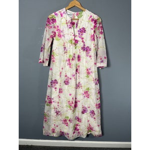 Puede incluir: Vestido maxi blanco con estampado floral en tonos rosa y morado. El vestido tiene un escote en V y mangas 3/4. La etiqueta "Ael's" es visible en el vestido.