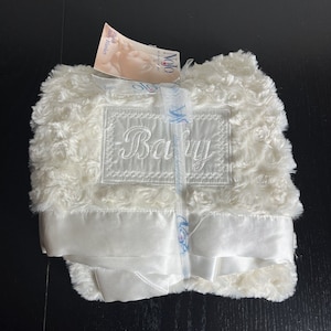 May include: White baby blanket with a satin trim and embroidered "Baby" in the center. The blanket is made of soft, fluffy fabric.