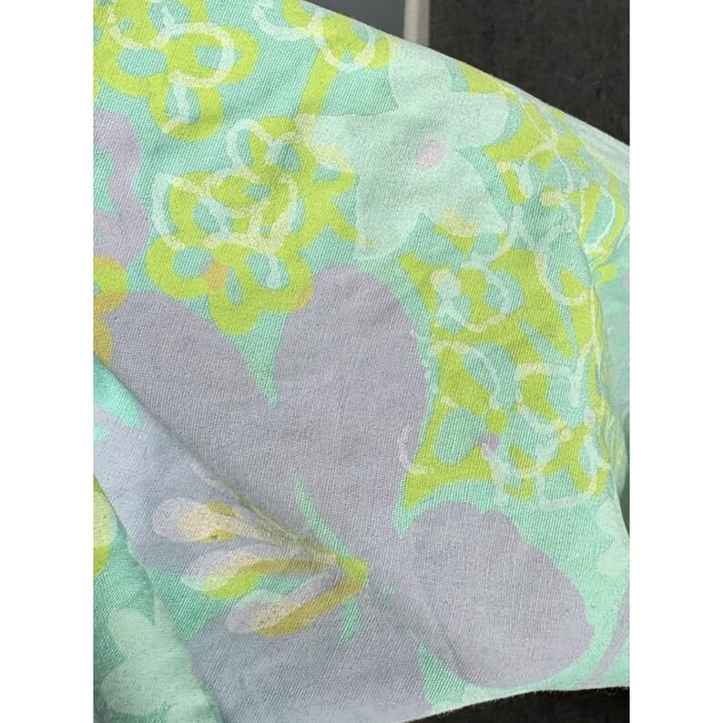 May include: Close-up of a fabric with a floral pattern. The design features large, light purple flowers and smaller, yellow and white flowers on a light green background. The fabric appears soft and slightly creased.