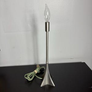 May include: A silver table lamp with a teardrop-shaped shade and a long, thin stem. The lamp has a clear light bulb and a cord with a plug.