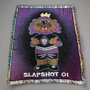 May include: Purple woven tapestry with a cartoon lion wearing a crown and a hockey jersey with the text "SLAPSHOT 01".