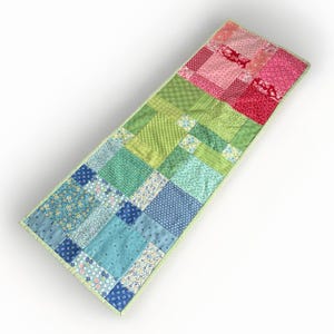 May include: A rectangular patchwork table runner with a variety of colorful fabric squares. The runner features a gradient of colors, transitioning from blue to green to pink and red. The edges are finished with a light green border.