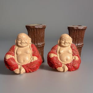 May include: Two smiling Buddha figurines in a seated position, wearing red robes, with a light beige skin tone. Two brown bamboo-style vases are behind the figurines. The background is a solid gray.