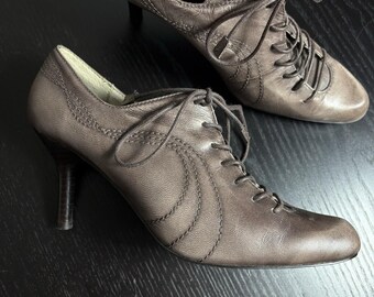 May include: A pair of brown leather lace-up heeled shoes. The shoes have a pointed toe and a small heel. The laces are tied in a bow. The shoes are on a dark surface.