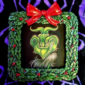 May include: A framed artwork featuring a green Grinch character with a mischievous grin. The frame is decorated with green holly leaves and red berries, topped with a glossy red bow. The artwork is set against a black background.
