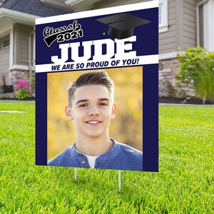 May include: A blue and white graduation yard sign with a photo of a young man. The sign says "Class of 2021 Jude We are so proud of you!"