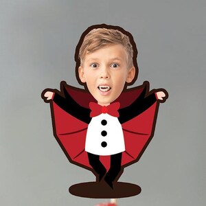 Puede incluir: Un cupcake cubierto con glaseado blanco y una frambuesa fresca. Una imagen de dibujos animados de un niño disfrazado de vampiro está encima del cupcake. El vampiro tiene una capa roja y una pajarita.