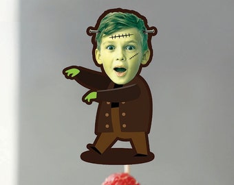 May include: A cupcake with white frosting and a raspberry on top. A green Frankenstein monster cupcake topper with a surprised expression is on the cupcake.